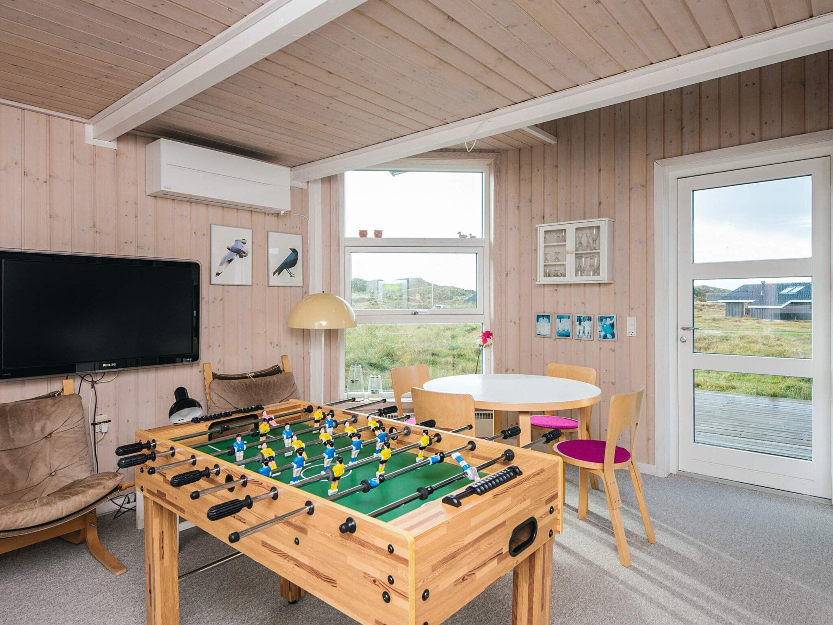 A room with a foosball table, a television, a table with chairs, and a window with a view of the outdoors.