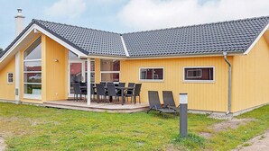A yellow building with a sloped roof, a covered patio with chairs, and a grassy area in front. - Holiday Home in Großenbrode (Grossenbrode)