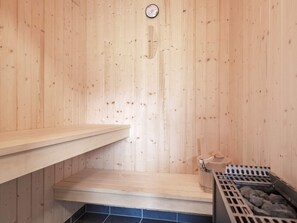 A sauna room with wooden walls, a wooden bench, a small table with a bottle, and a heater with rocks.
