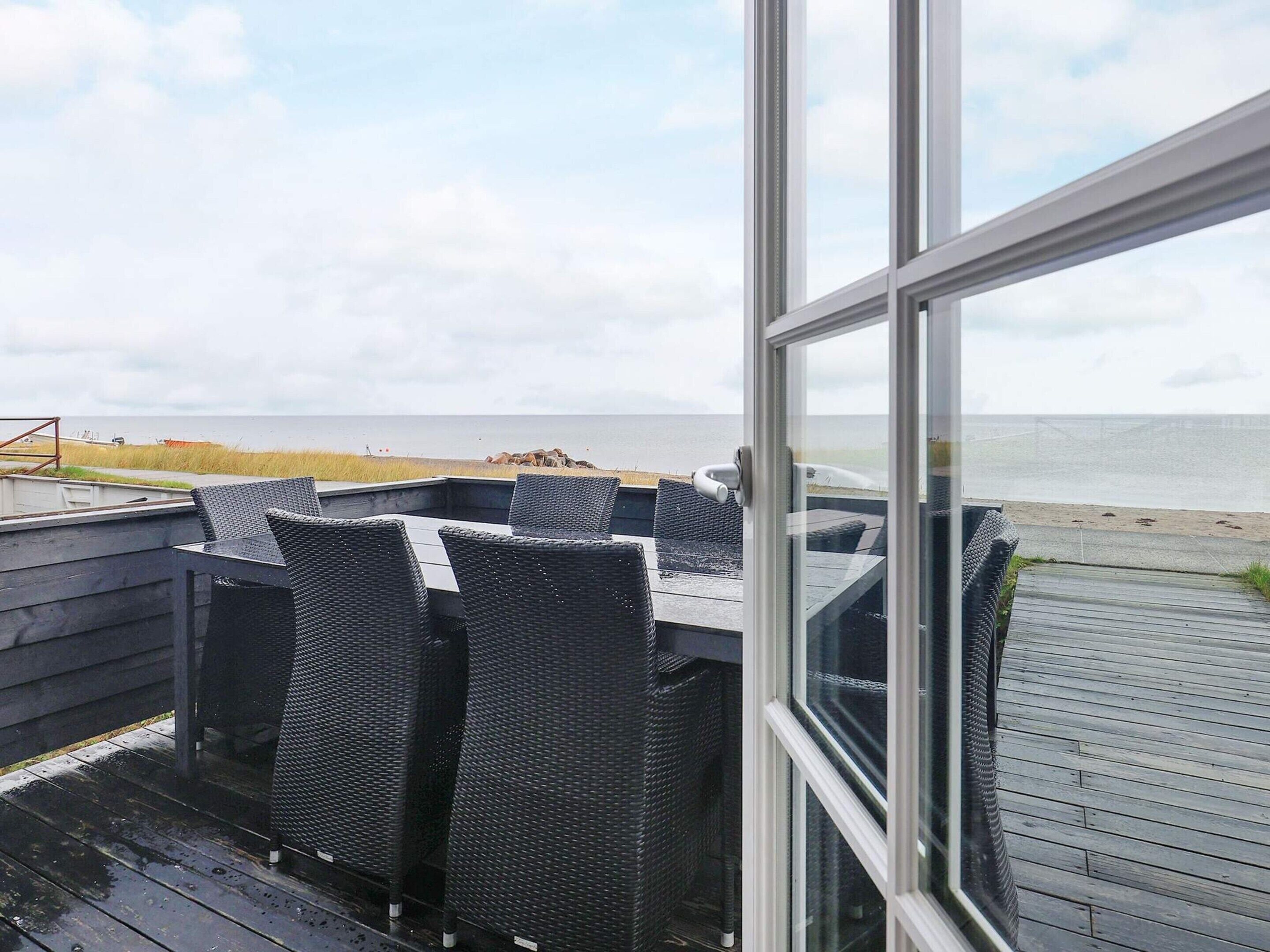 Une vue depuis une fenêtre donnant sur une plage, avec une terrasse en bois et des meubles en osier.