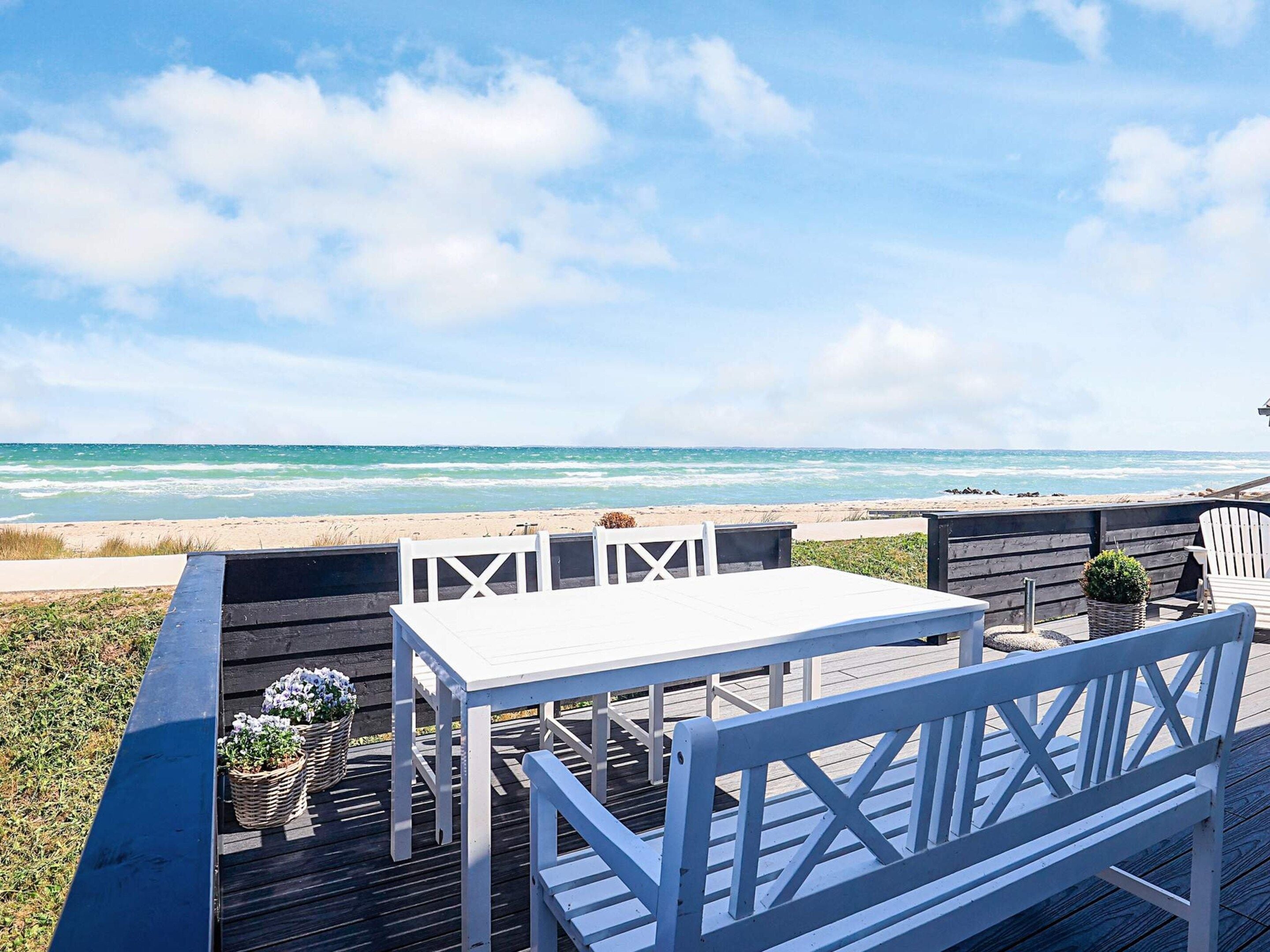 A wooden deck with a white table and bench, overlooking a beach with waves.
