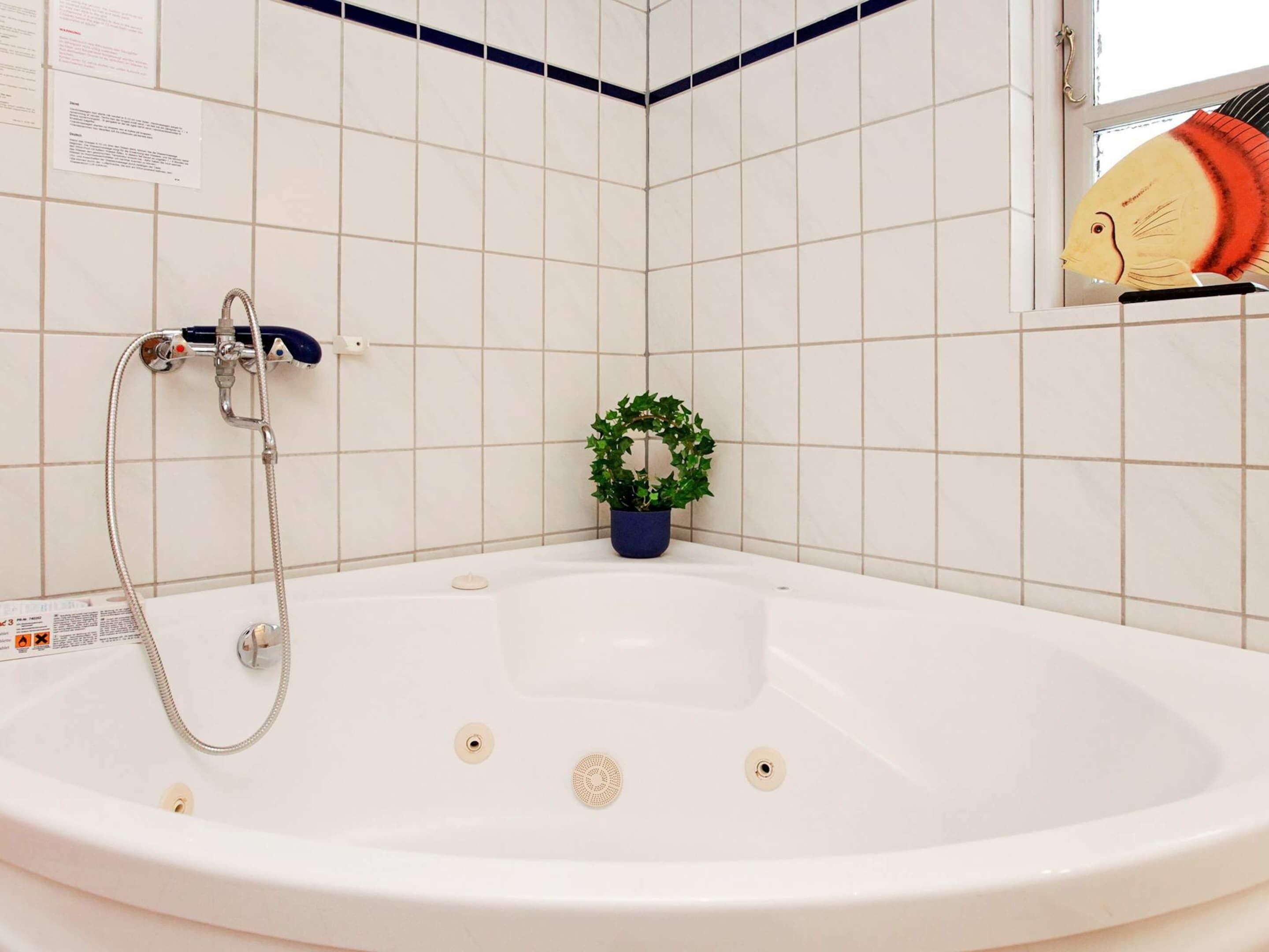Et badeværelse med badekar, bruser, en potteplante og et vindue med en pynteting.
