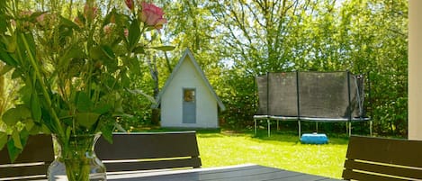 Un área de estar al aire libre con una mesa, sillas, un pequeño edificio blanco y un trampolín al fondo.