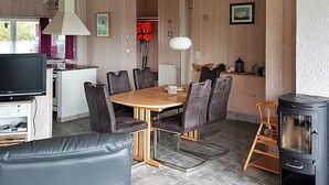 A cozy living room with a fireplace, a dining area with a wooden table and chairs, and a kitchen area with a television and a red life jacket hanging on the wall. - Holiday Home in Otterndorf (Otterndorf)