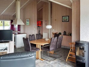 A cozy living room with a fireplace, a dining area with a wooden table and chairs, and a kitchen area with a television and a red life jacket hanging on the wall. - Holiday Home in Otterndorf (Otterndorf)