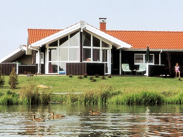 Une maison avec un toit en pente, de grandes fenêtres et un étang où nagent des canards.