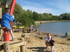 A sandy beach area with wooden structures, including a swing set and tables, near a body of water. - Holiday Home in Otterndorf (Otterndorf)