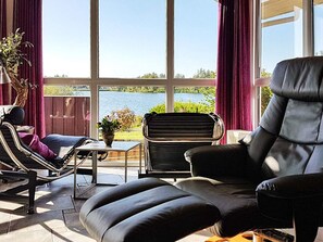 A sunlit room with a view of a lake, featuring a sofa, a chair, and a small table with a plant. - Holiday Home in Otterndorf (Otterndorf)