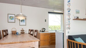 Et hyggeligt køkken med træskabe, et spisebord og stole. Der er et vindue med udsigt til grønne områder, en hængende lampe og en hylde med dekorative genstande.
