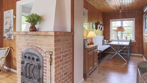 A cozy cabin interior with a brick fireplace, wooden walls, a dining area with a table and chairs, and a window with a view. - 6 Person Holiday Home in Gilleleje (Gilleleje)