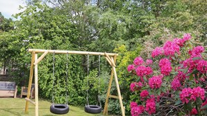 A wooden swing set with two swings, a grassy lawn, and a bush with pink flowers. - 6 Person Holiday Home in Gilleleje (Gilleleje)