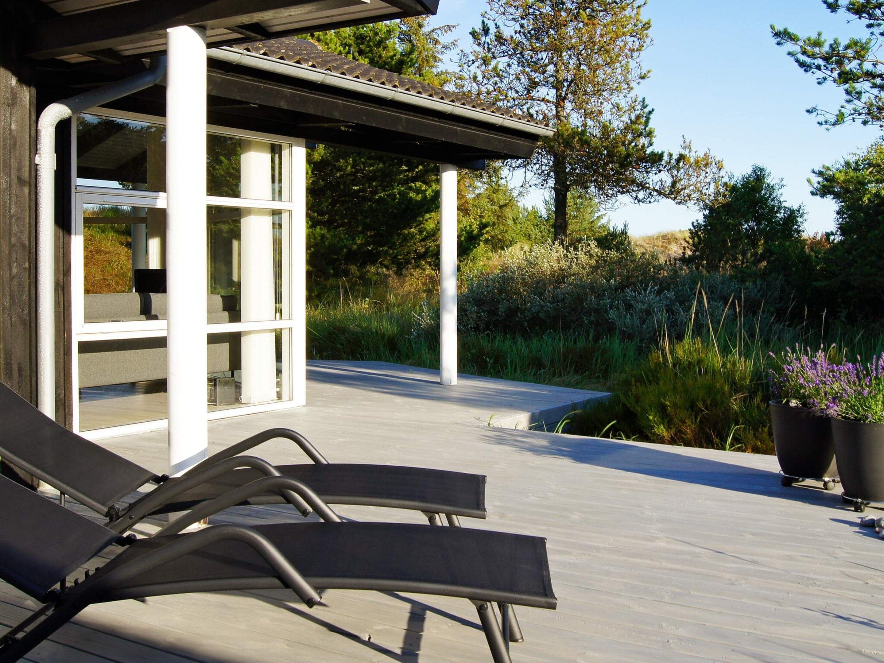 A modern outdoor deck with lounge chairs, a glass-enclosed structure, and a garden with tall grasses and trees.