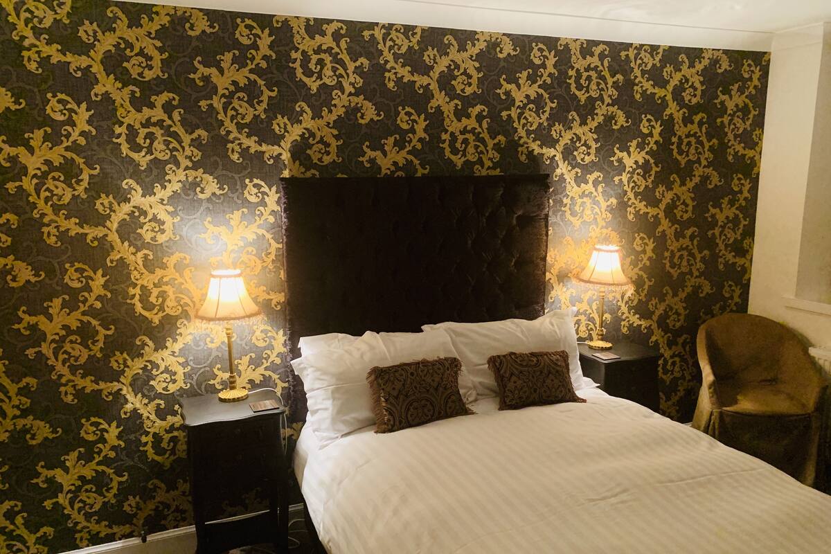 A bedroom with a large bed, two bedside tables with lamps, a chair, and a patterned wallpaper.
