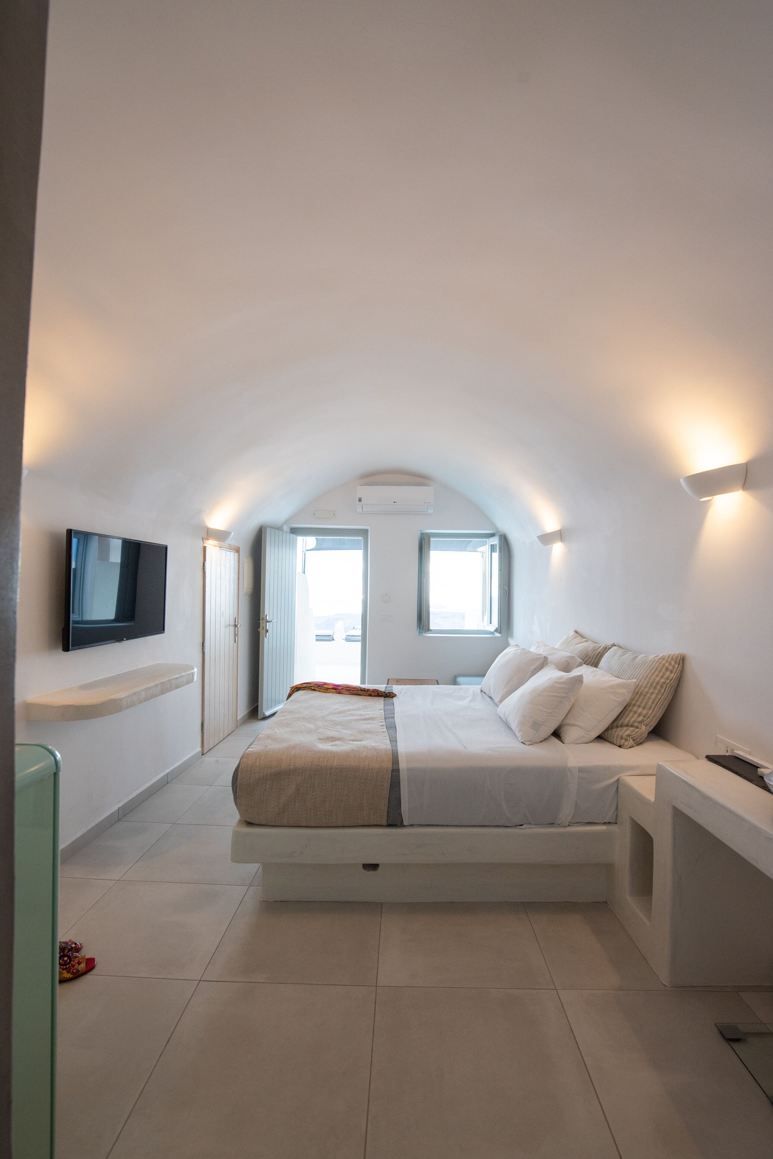 A modern bedroom with a bed, a TV mounted on the wall, and a window providing natural light.