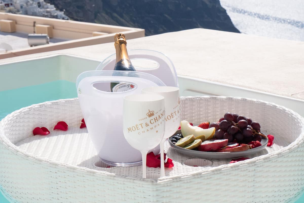 A white wicker tray with a bottle of champagne, two glasses, and a plate of fruit on a poolside deck with a view of the ocean and cliffs.