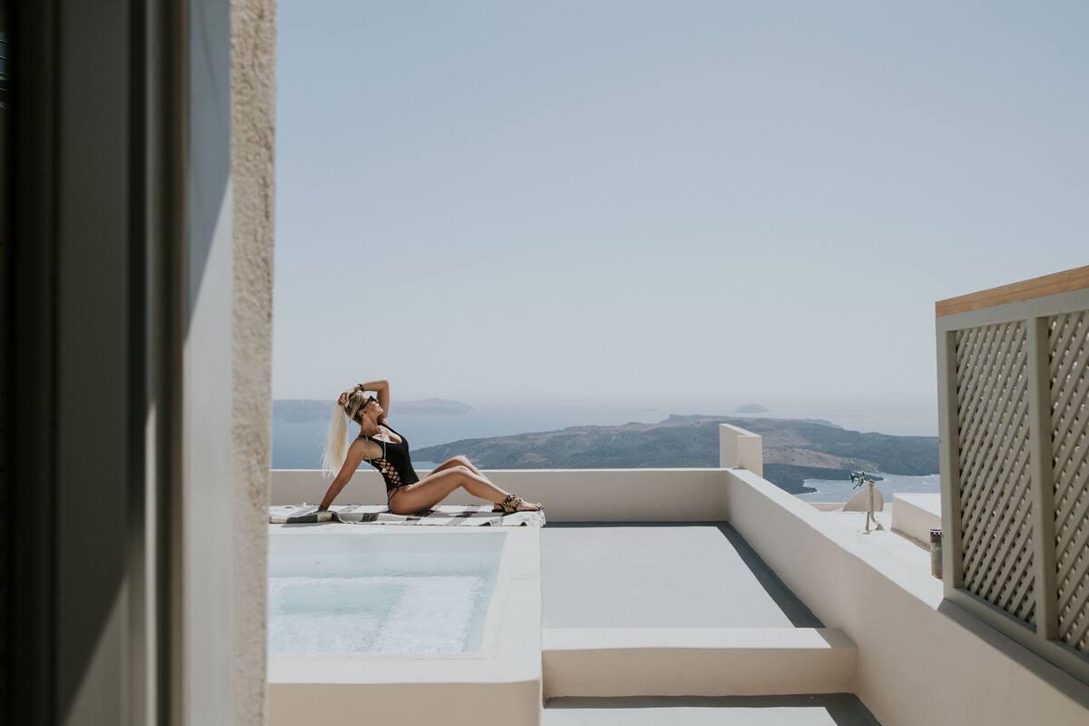 A woman sitting on a balcony with a pool, overlooking a scenic view of hills and the sea.