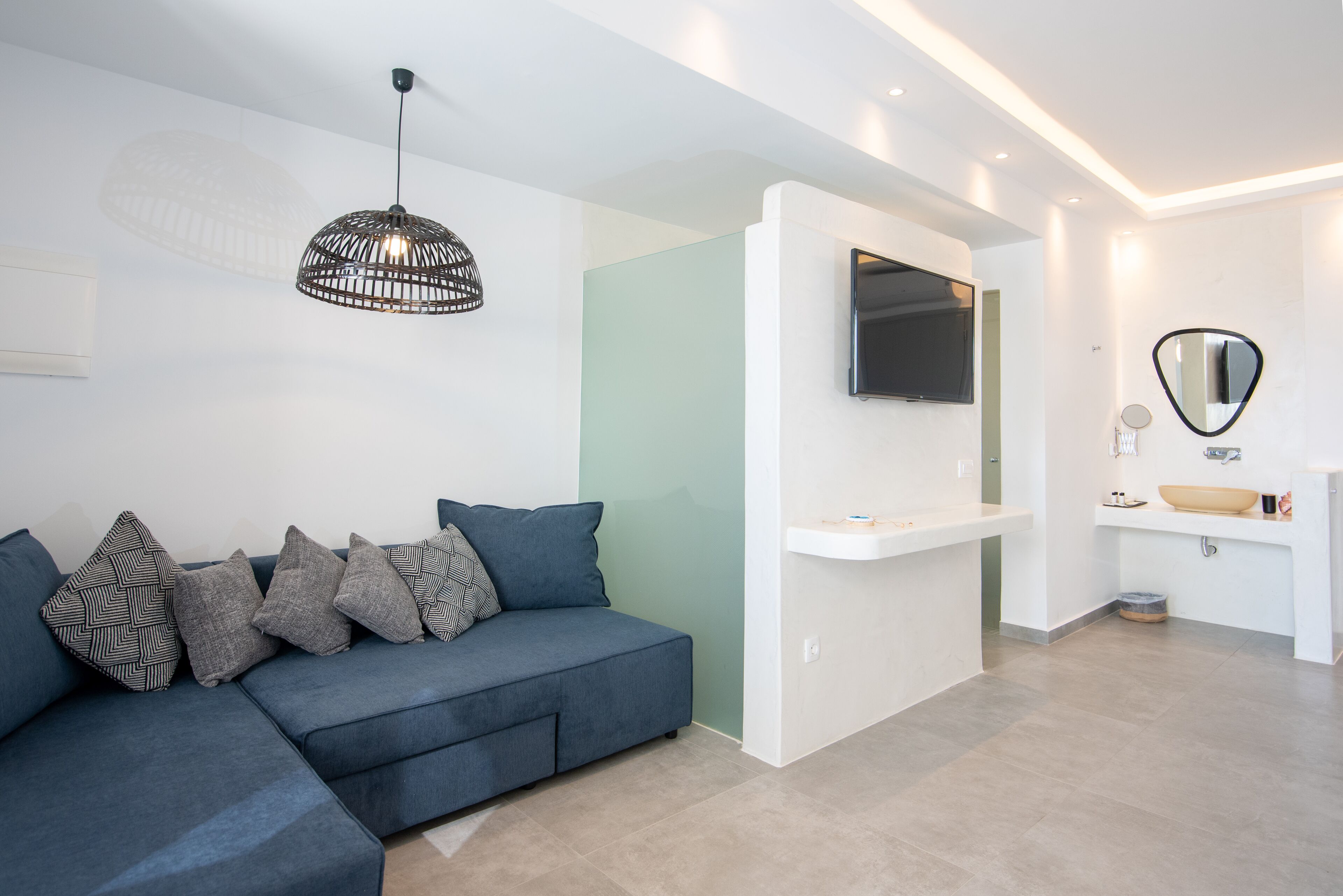 A modern living room with a sectional sofa, a hanging light fixture, a mounted TV, and a minimalist bathroom with a sink and mirror.