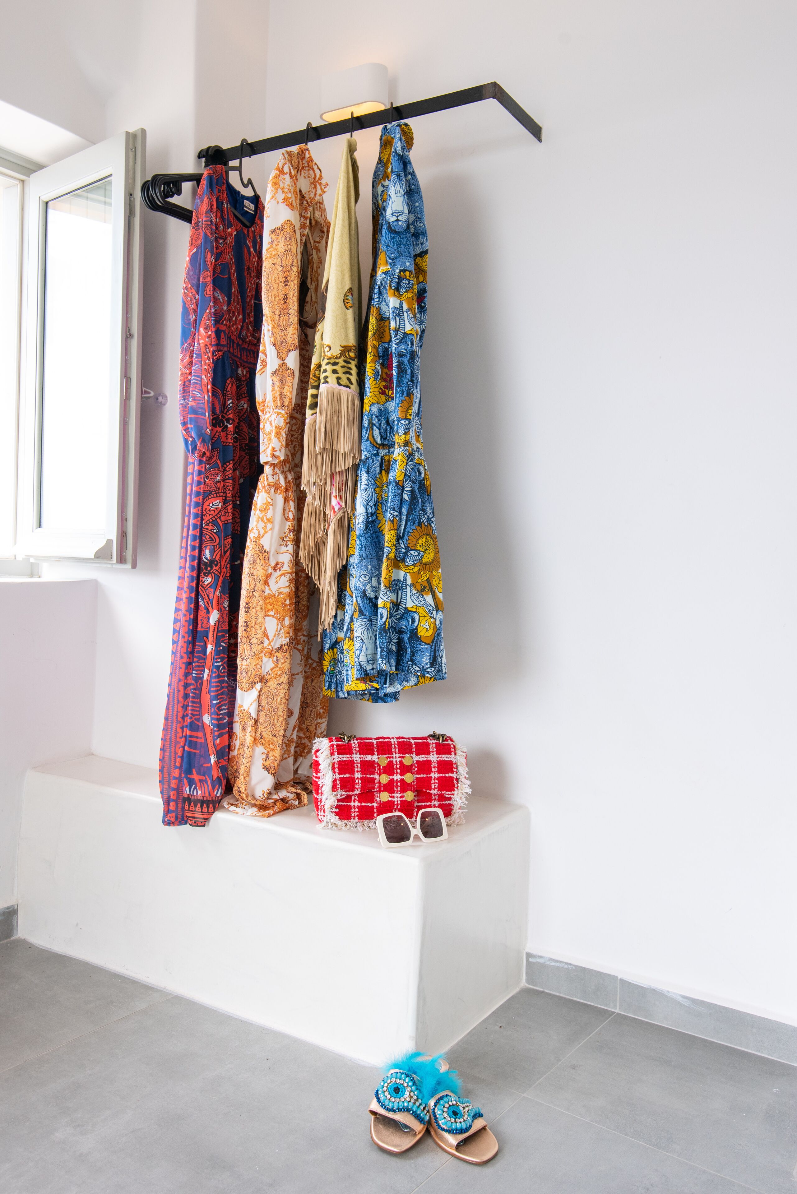 Ein Zimmer mit Fenster, ein Kleiderständer mit verschiedenen Kleidungsstücken, eine rote, karierte Tasche und ein Paar blaue Sandalen auf dem Boden.