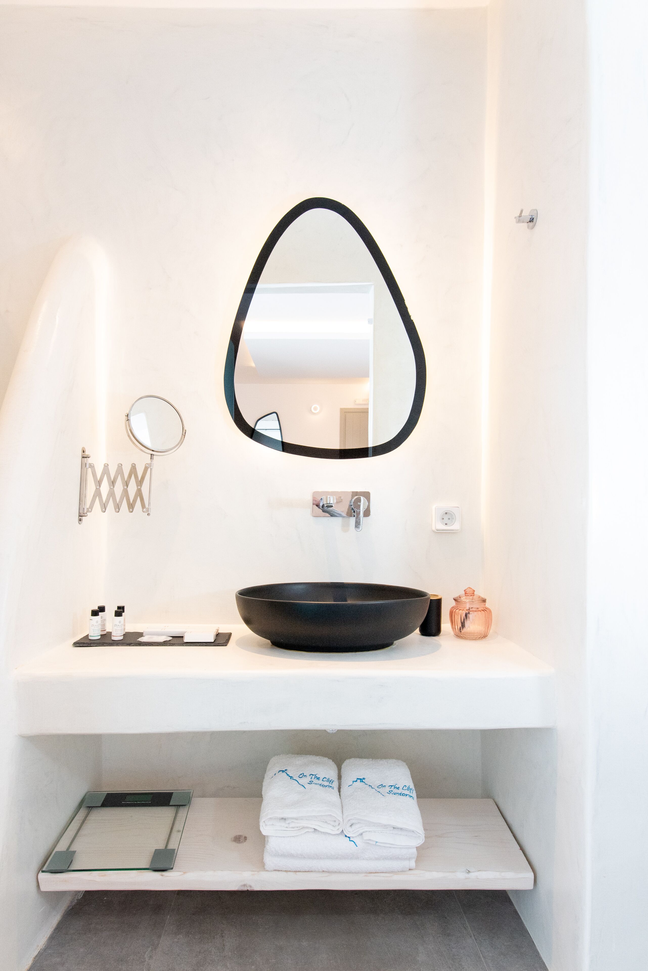A bathroom with a black oval mirror, a round mirror, a black basin, and a shelf with folded towels.