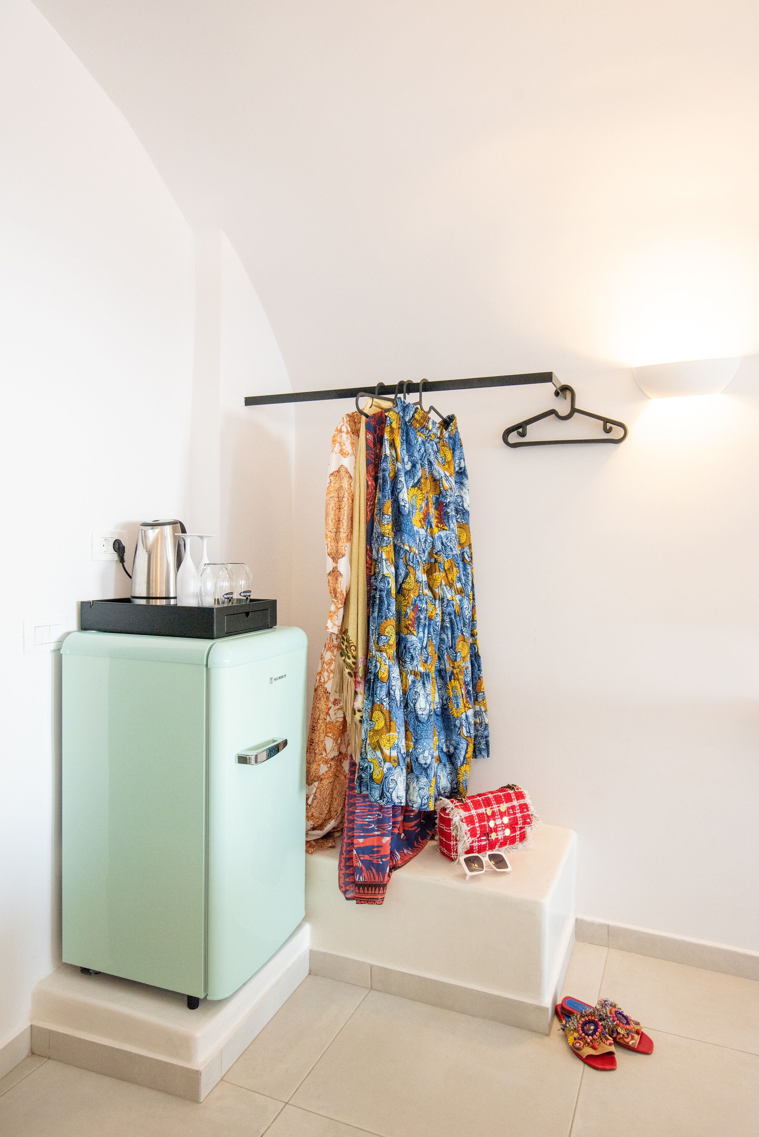 A compact room with a vintage-style refrigerator, a small table with a kettle and glasses, a hanging coat rack, and a pair of slippers on the floor.
