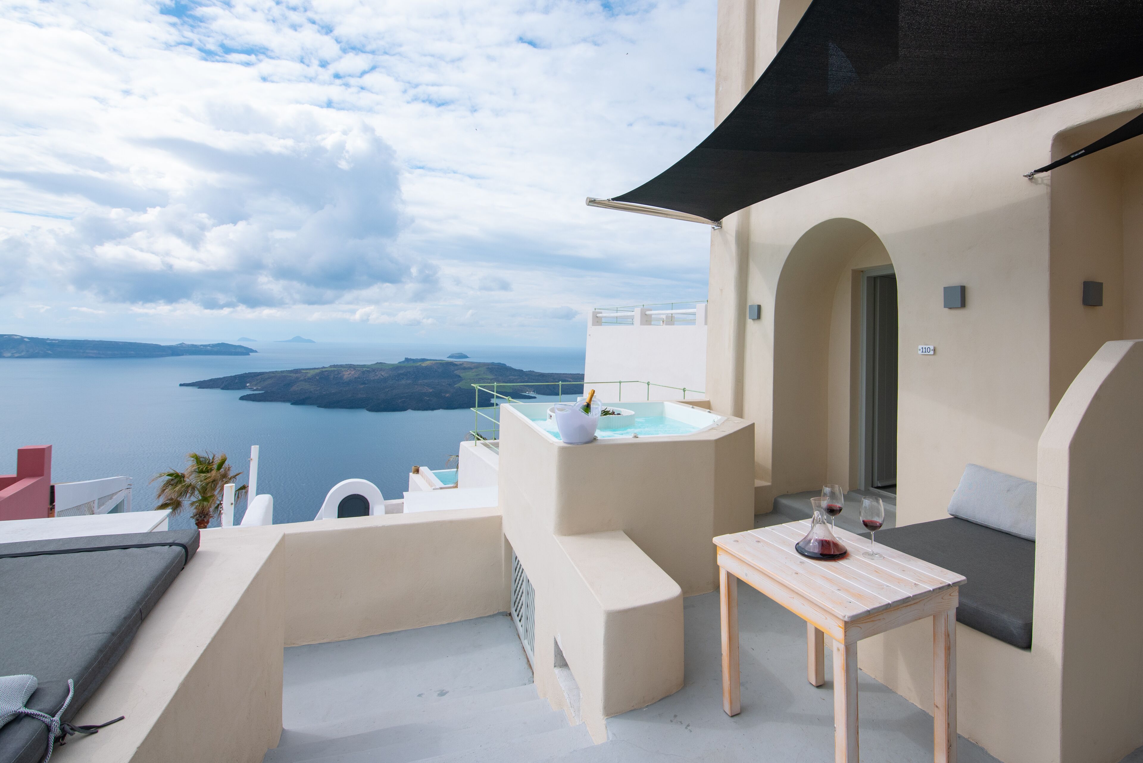 A balcony with a view of the sea, a hot tub, and a table with wine glasses.