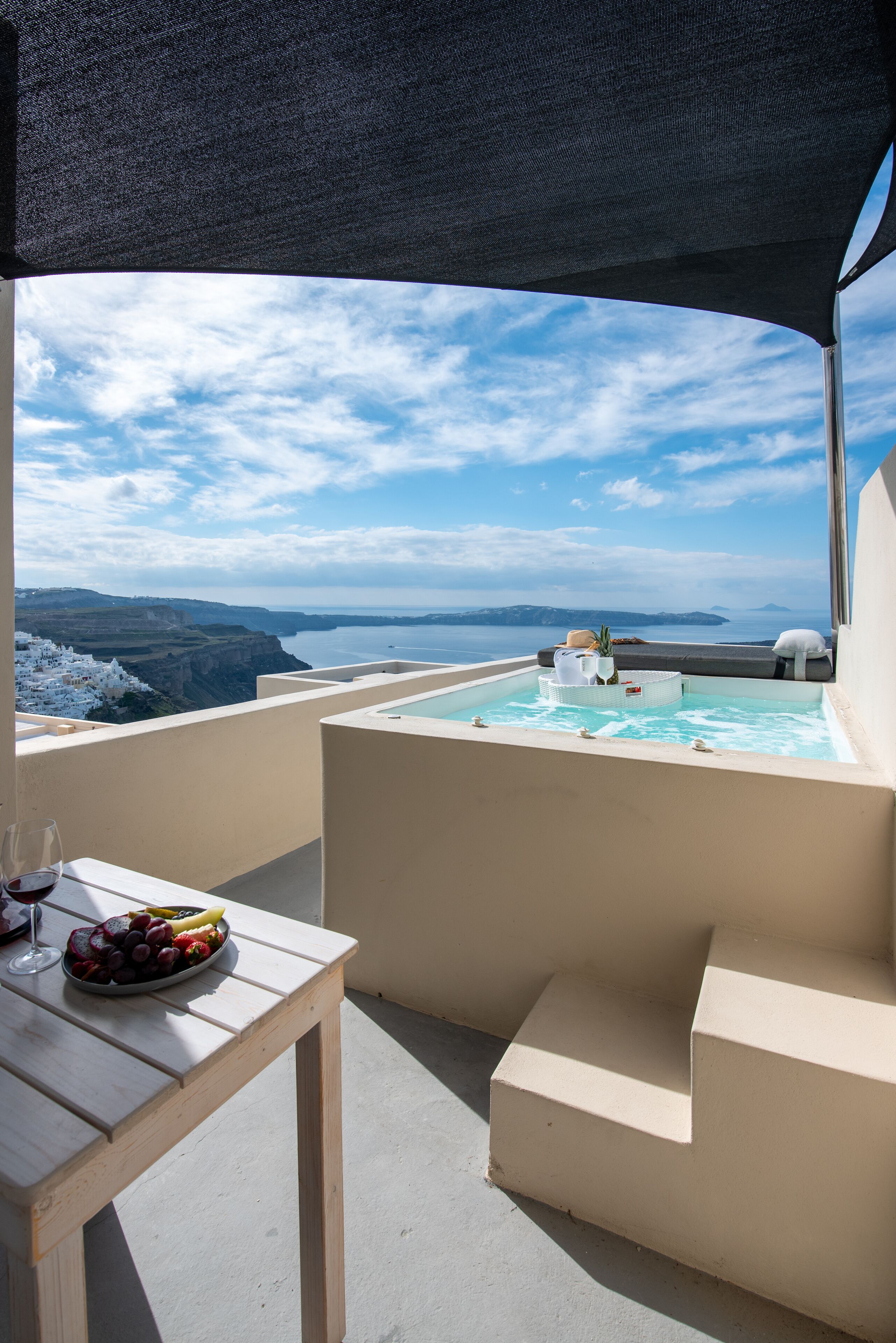 A terrace with a pool, a table with fruit and wine, and a view of the sea and cliffs.