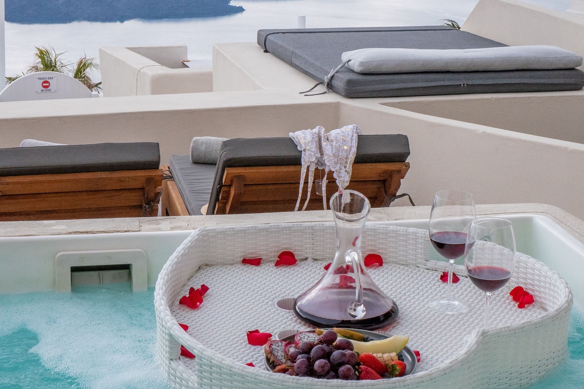 A hot tub with a tray of fruits and wine glasses, surrounded by lounge chairs and a scenic view of the sea.