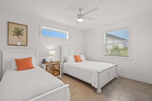 Un dormitorio con dos camas, un ventilador de techo, una mesita de noche con lámpara y una ventana con persianas.