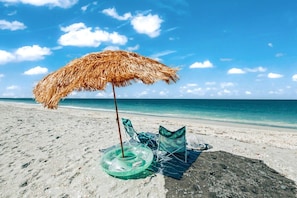 Ein Strand mit einem Strohdachschirm, einem grünen Schwimmreifen und zwei Liegestühlen.