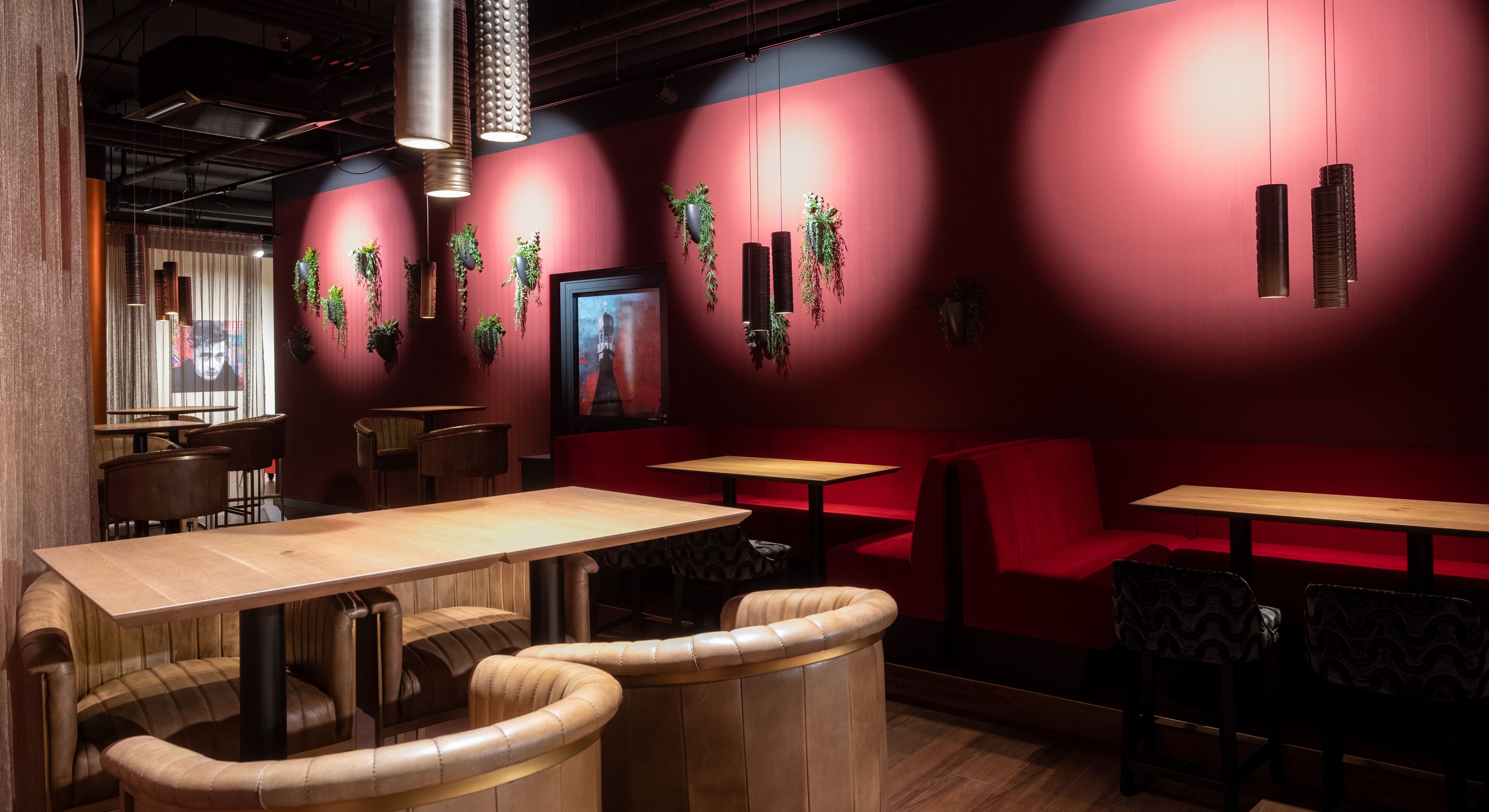 Ein Restaurantinnenraum mit Holztischen und -sitzen, rot gepolsterten Bänken, hängenden Pflanzen und einem gerahmten Bild an der Wand.