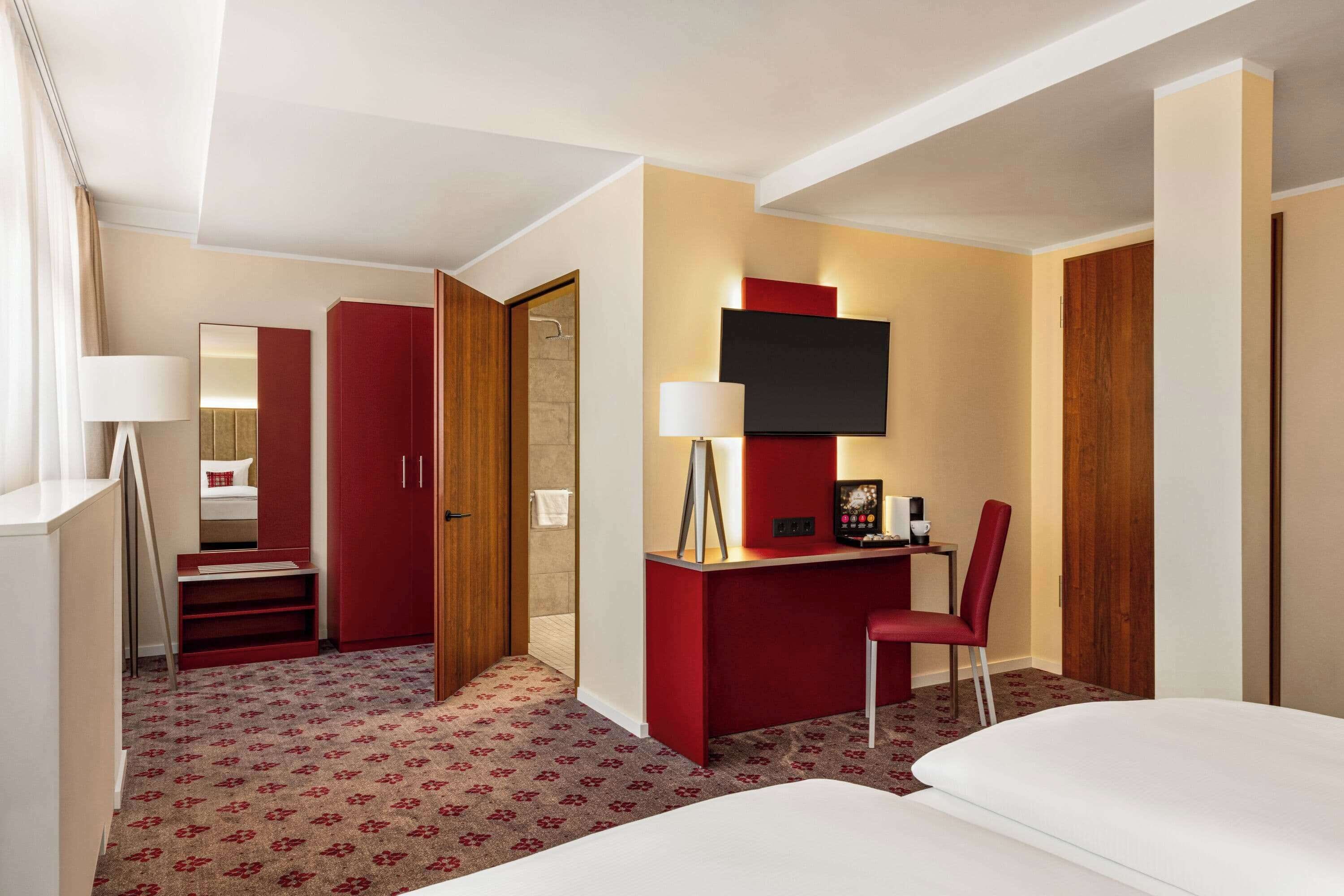 Una habitación de hotel con cama, escritorio con silla, televisor, espejo y armario.