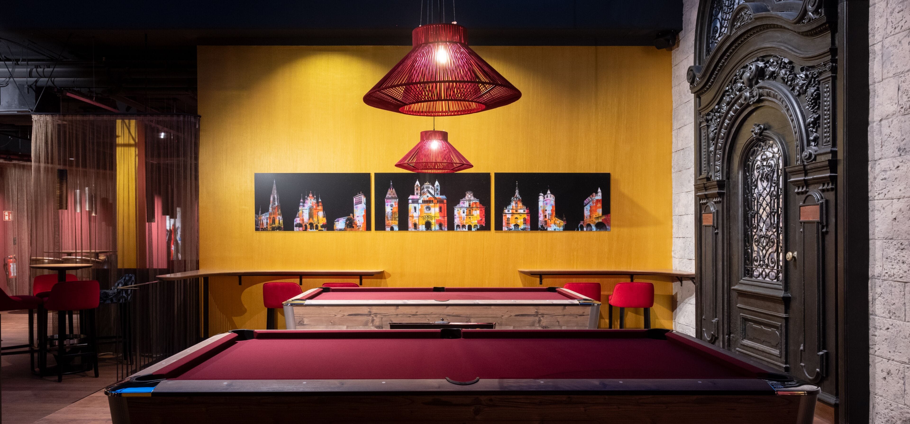 A billiards table with red cushions, a red pendant light, and three paintings of buildings on a yellow wall.