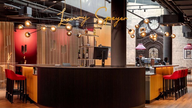A bar area with a curved counter, multiple stools, and hanging pendant lights. - Hotel Postgalerie Speyer (Speyer)