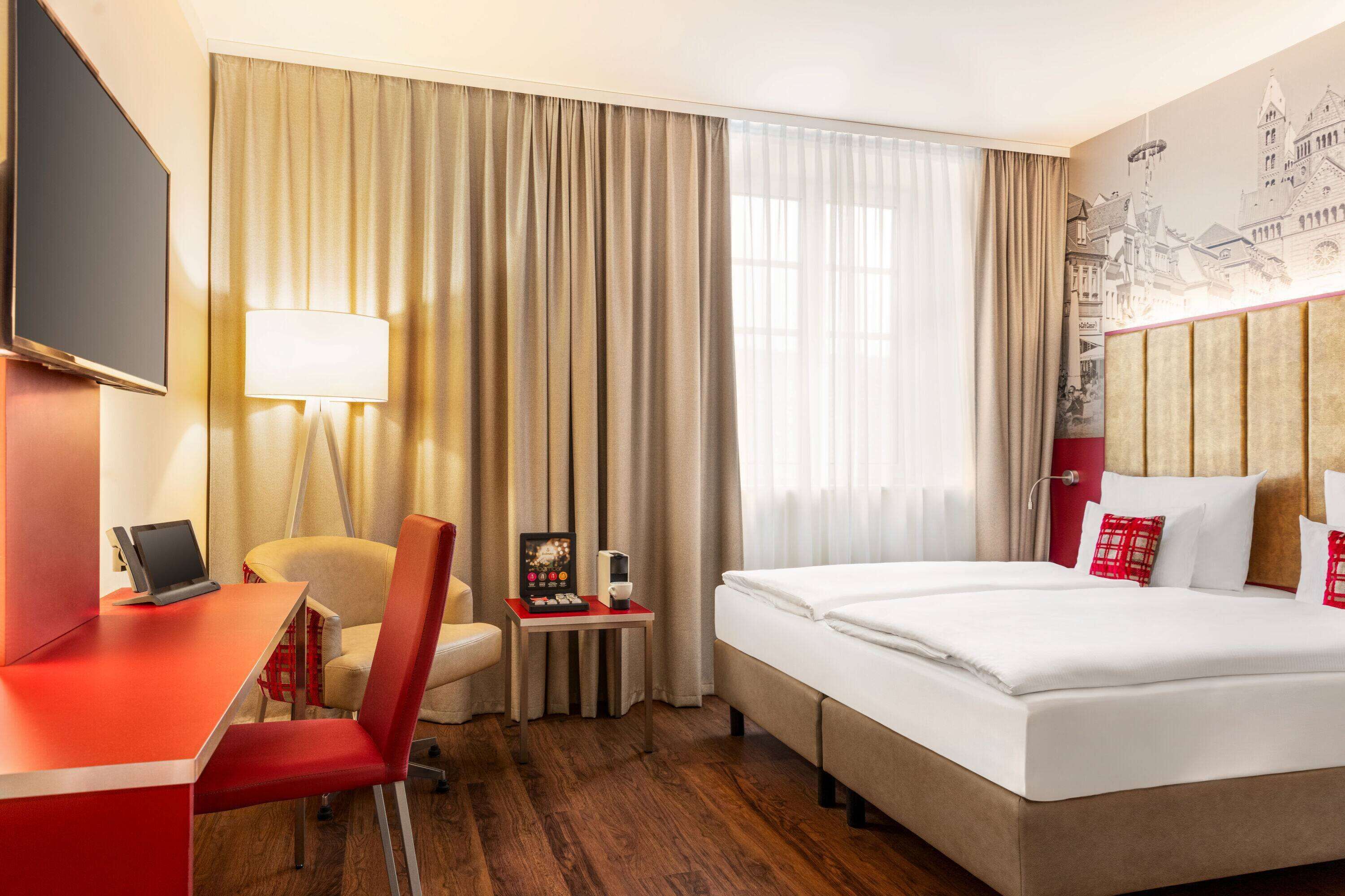 A hotel room with a bed, a desk with a laptop, a chair, a small table with a lamp, and a wall with a cityscape mural.