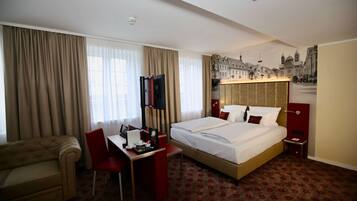 A hotel room with a bed, a desk, a chair, a TV, and a cityscape mural on the wall.