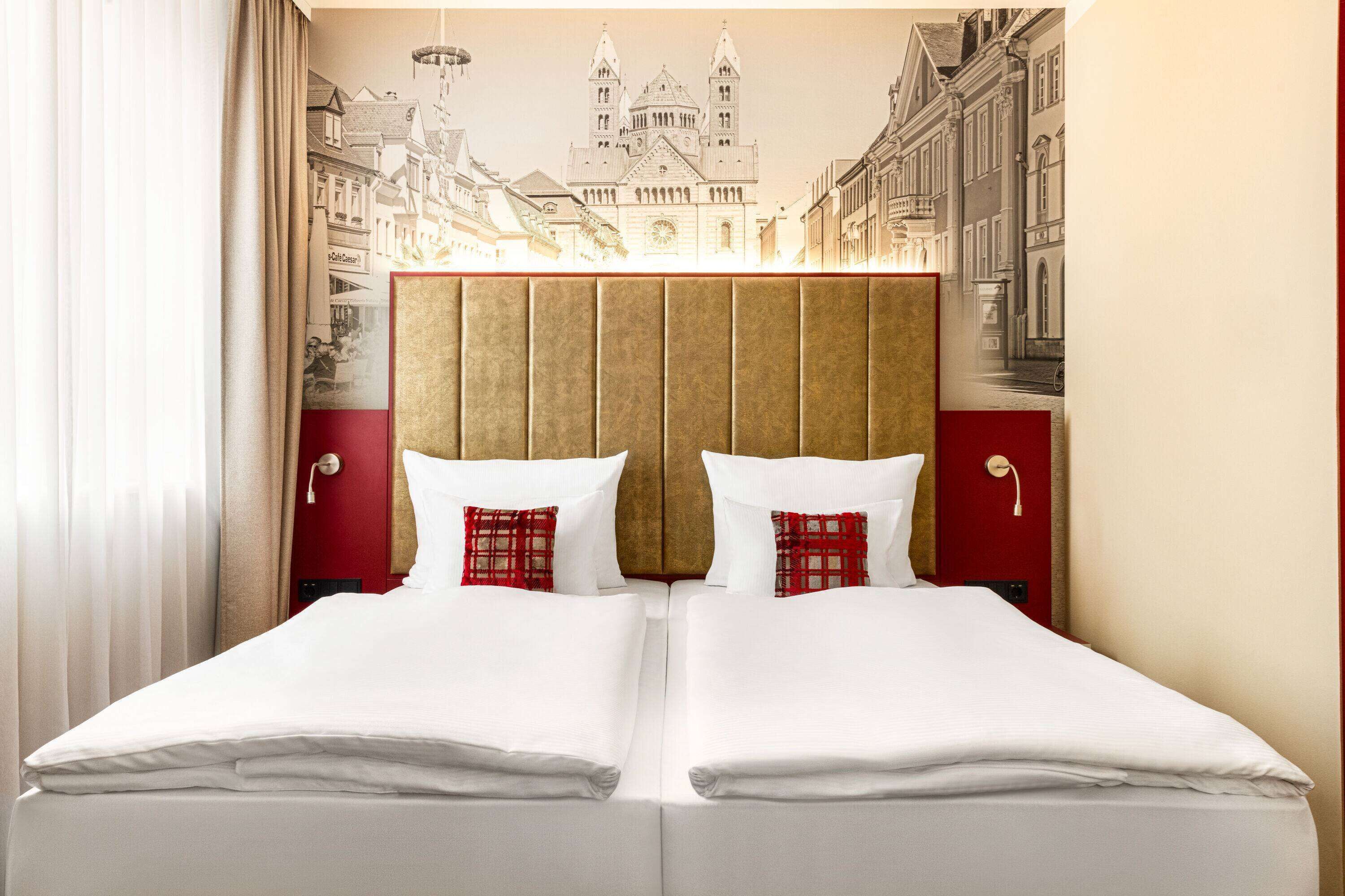 A double bed with white linens and red plaid pillows, a wooden headboard, and a mural of a cityscape on the wall.