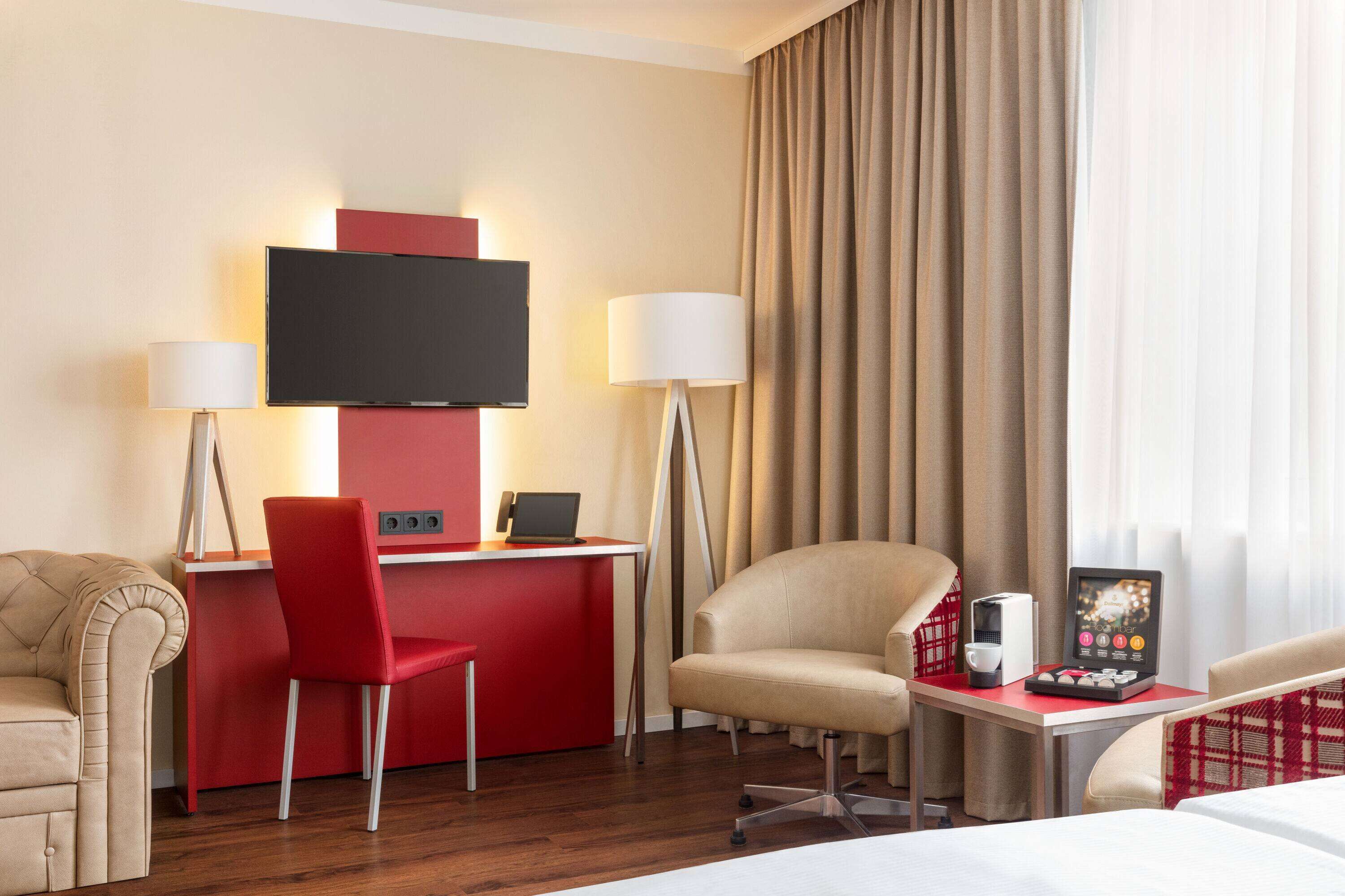 A hotel room with a desk, a television, a red chair, a beige armchair, a bed, and a window with curtains.