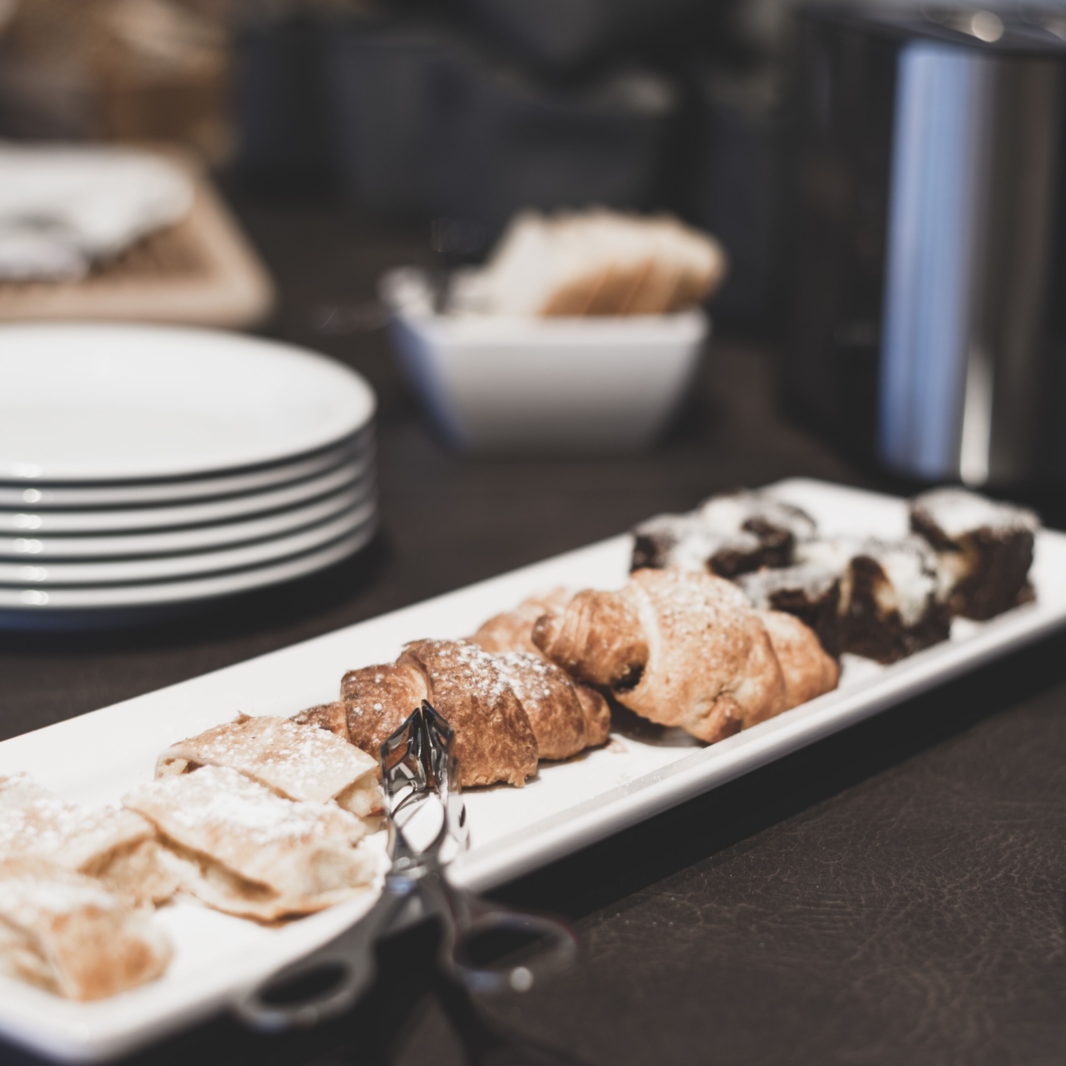 Une assiette rectangulaire blanche, sur laquelle sont disposées diverses pâtisseries, et une paire de ciseaux, le tout posé sur une surface sombre.