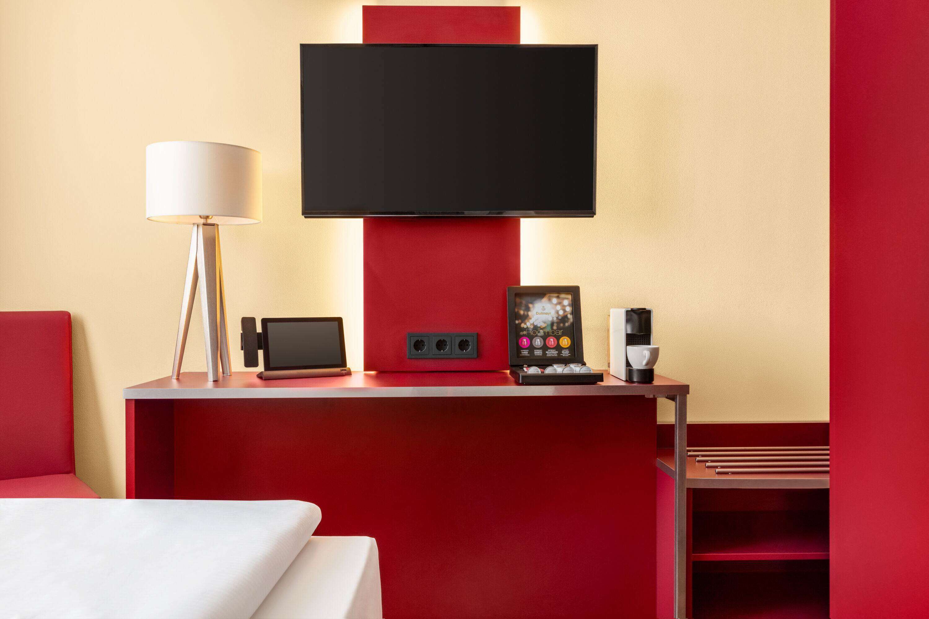 A hotel room with a red accent wall, a flat-screen TV mounted above a desk, a lamp, a coffee maker, and a small table with a picture frame.