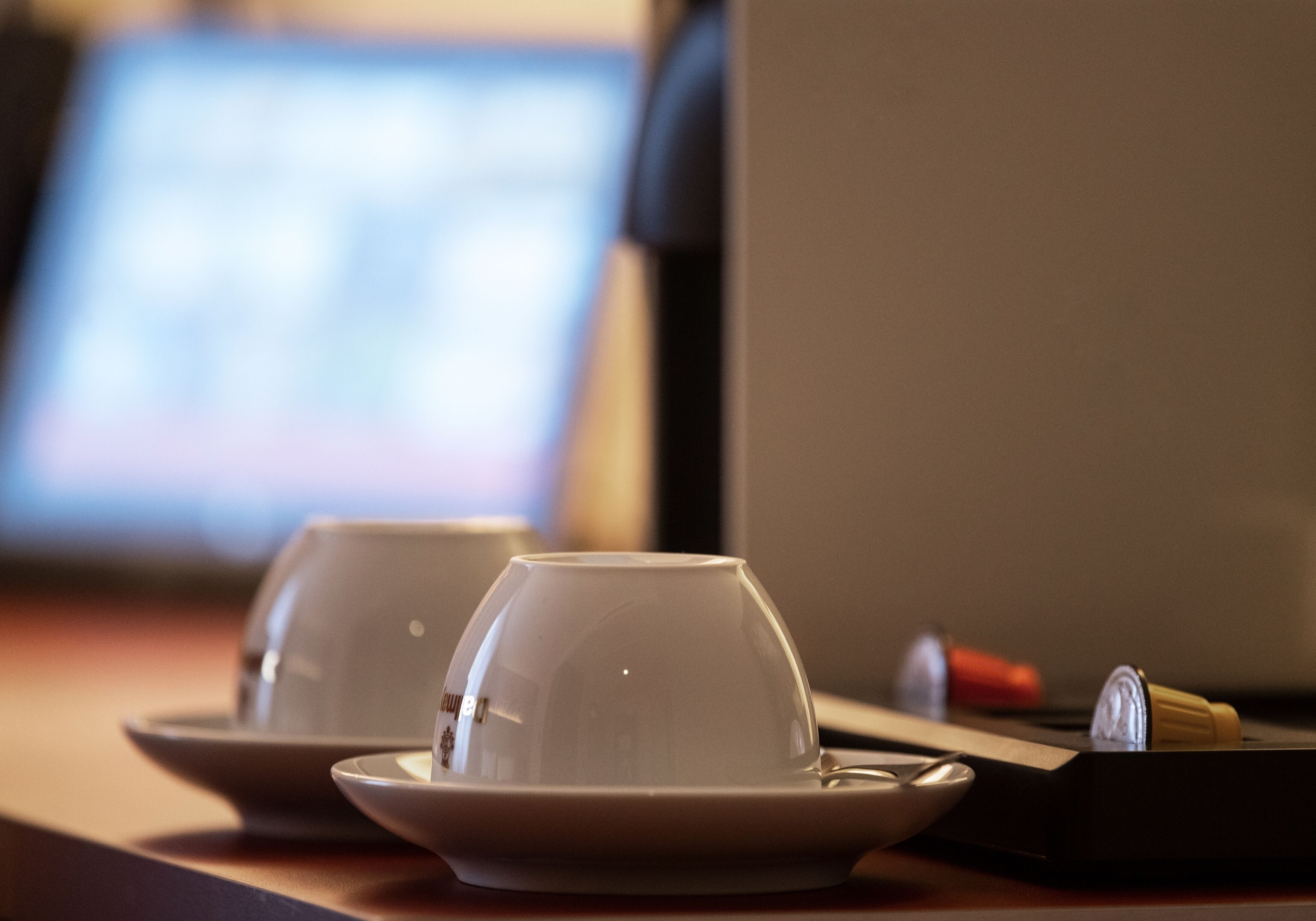 Two white cups on saucers on a table with a laptop and a red object in the background.