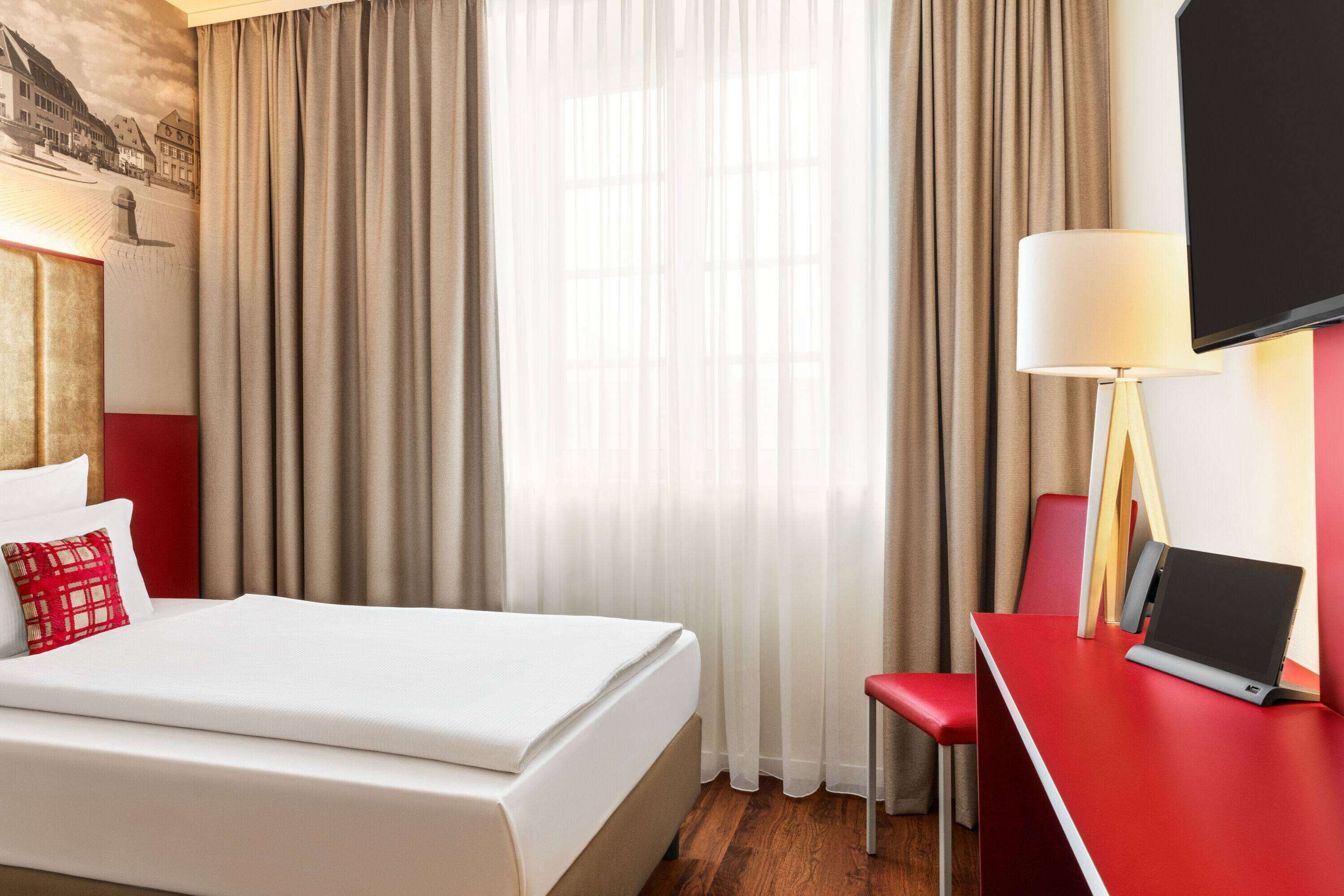 Ein Hotelzimmer mit einem Bett, einem Schreibtisch mit einem Laptop, einem Sessel und einem an der Wand montierten Fernseher.