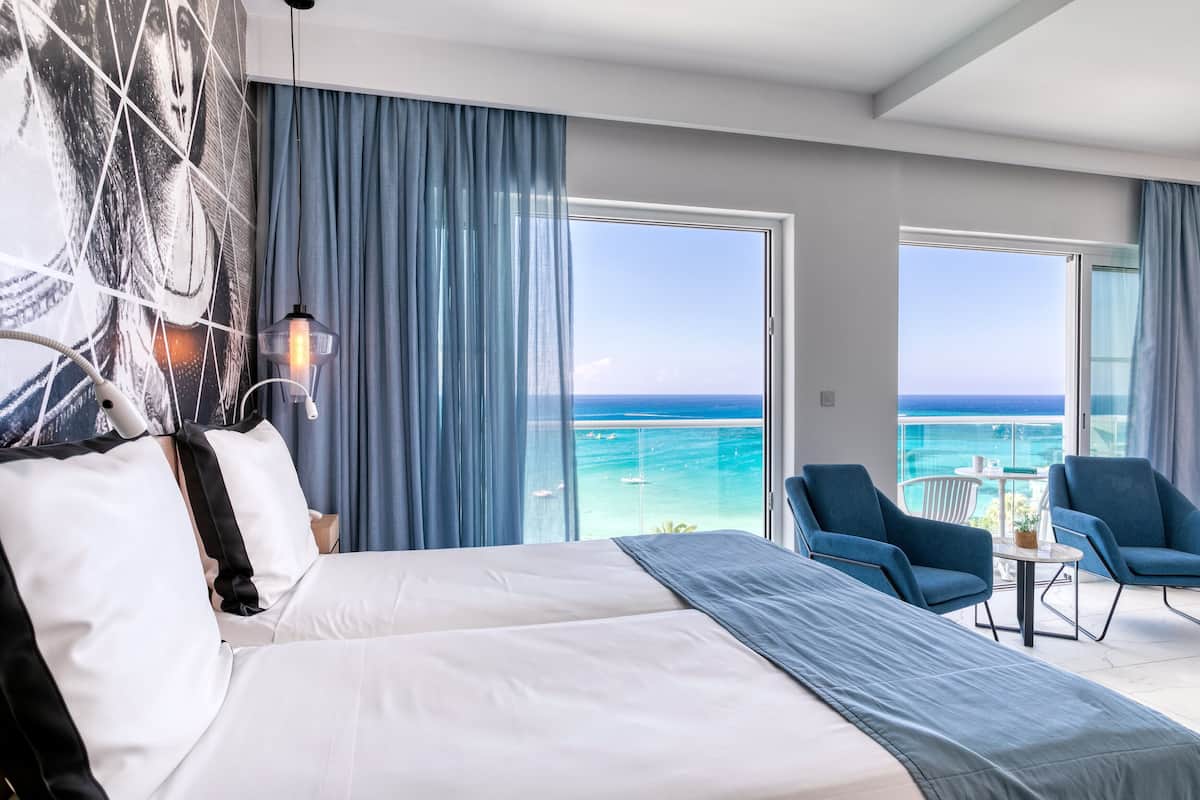 A hotel room with a large bed, a view of the ocean, and a balcony with chairs.