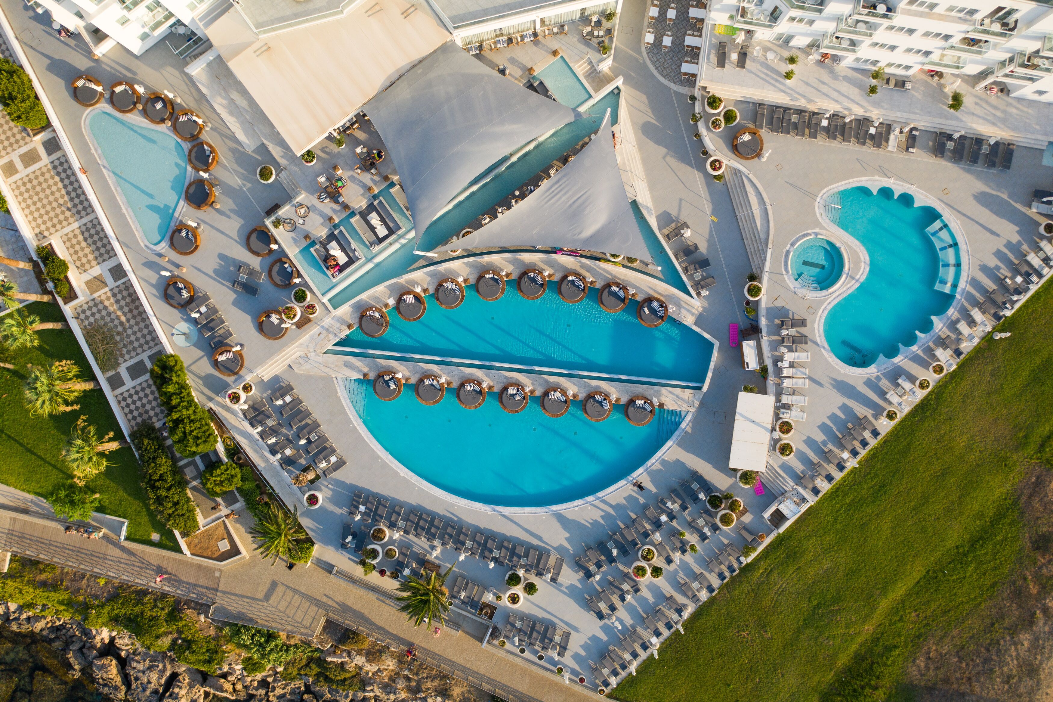 Vista aérea de un complejo turístico con múltiples piscinas, tumbonas y una plaza central.