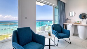 A modern hotel room with a balcony overlooking a beach, a round table, two blue armchairs, a white sink, and a mirror. - King Jason Protaras - Designed for Adults By Louis Hotels (Paralimni)