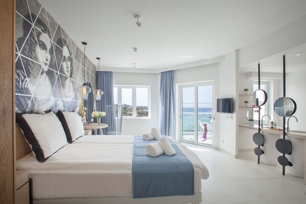 A modern hotel room with a large bed, a balcony with a view of the sea, a TV, and a bathroom with a mirror and sink.