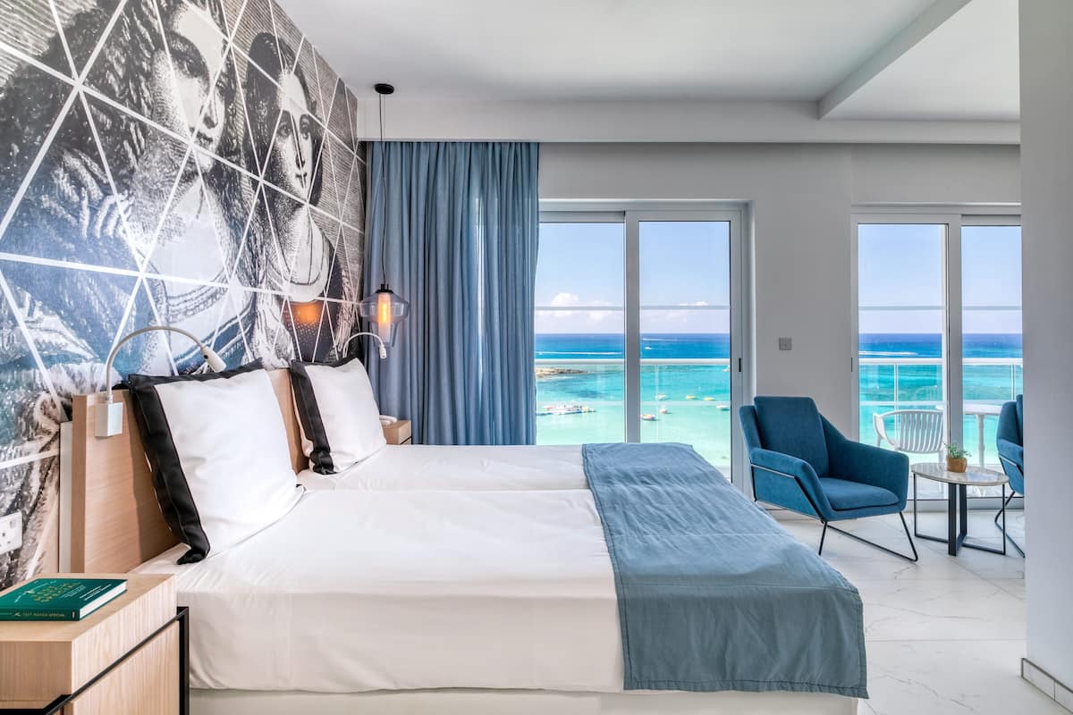 A modern hotel room with a large bed, a view of the ocean, a balcony with seating, and a geometric wall mural.