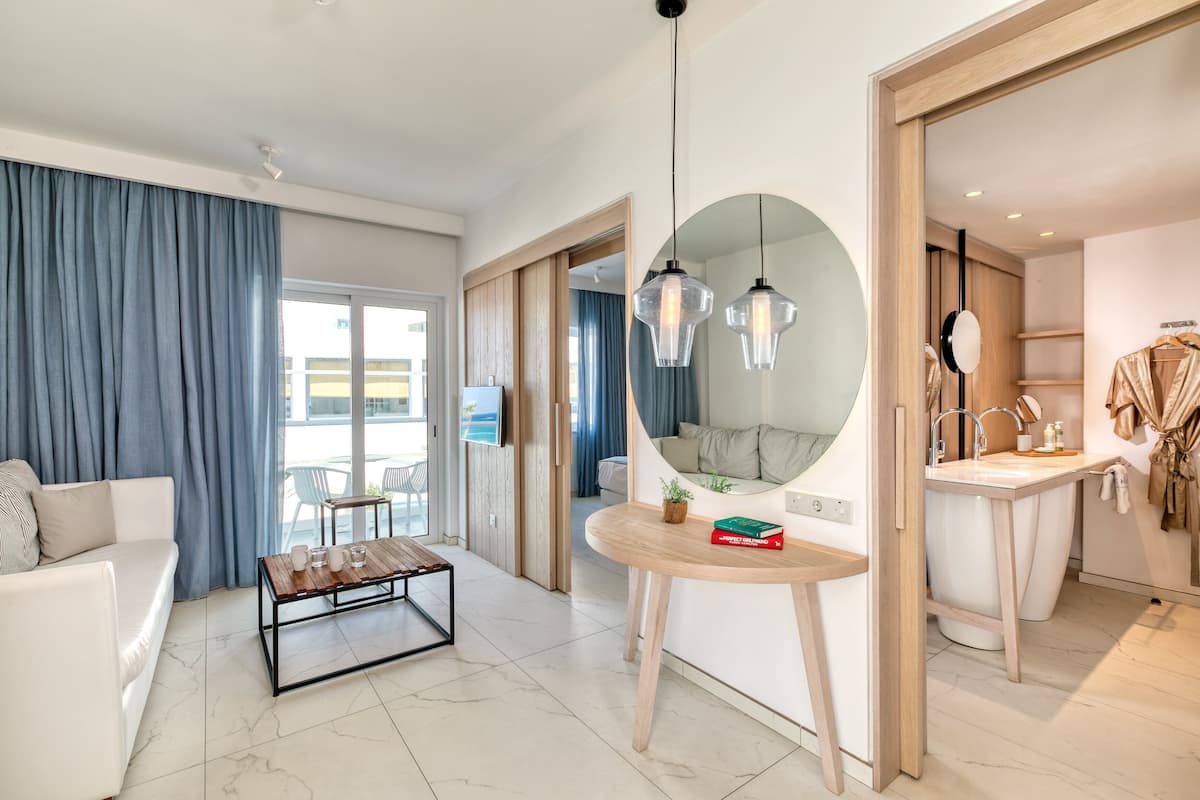 A modern hotel room with a living area, a dining table, a round mirror, a small round table with books, and a bathroom with a sink and towel rack.