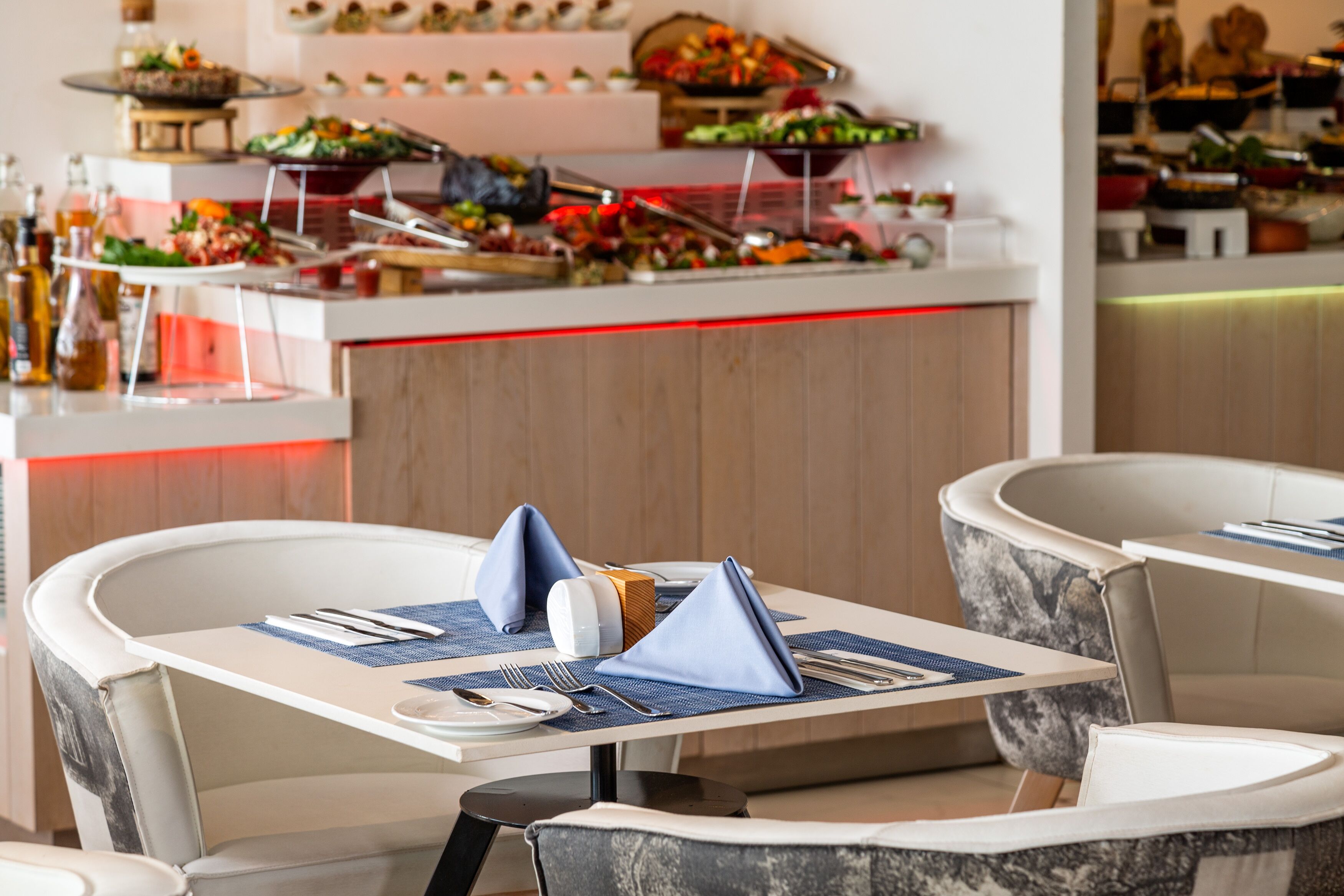 Un ambiente de restaurante con una zona de bufé al fondo, mesas ordenadas con servilletas azules y una variedad de alimentos presentados en el bufé.