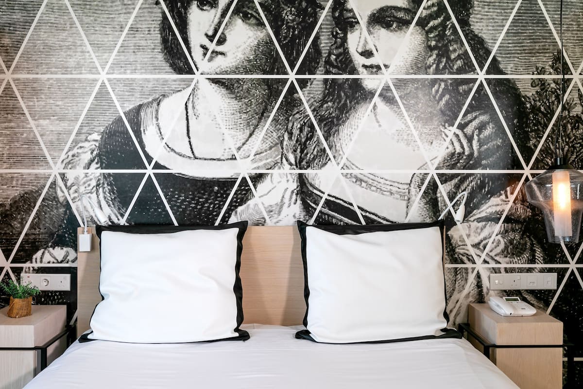A hotel room with a bed, two white pillows, a nightstand with a phone, and a wall with a geometric pattern.