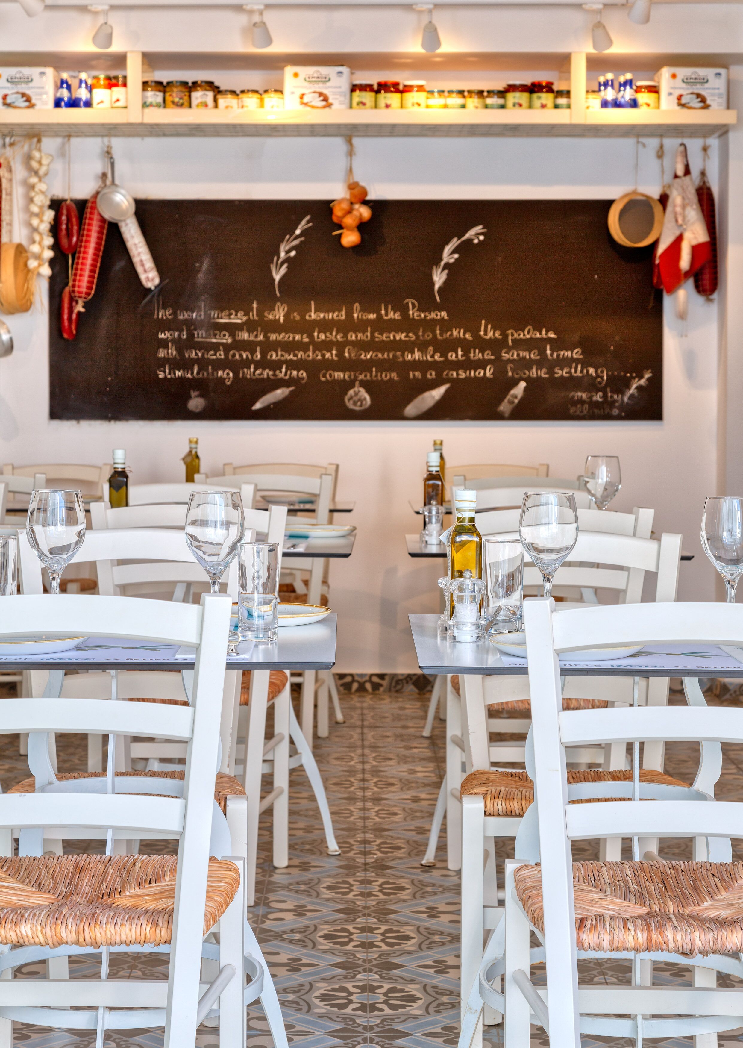 A restaurant interior with white tables and chairs, a chalkboard menu, and various condiments on shelves.